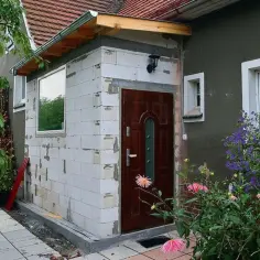 Budowa nowego pokoju do domu: białe pustaki, brązowe drzwi, okno i zielone rośliny.