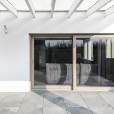Nowoczesne okna dąb bielony realizacje: przeszklone drzwi tarasowe z widokiem na wygodną sofę, biała ściana i pergola.