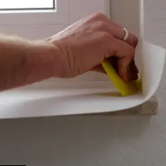 Dłoń z żółtą szpachelką przykleja białą okleinę na parapet. Poradnik: jak przykleić okleinę na parapet.
