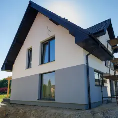 Nowy dom z dwukolorową elewacją, biało-szarą, w trakcie budowy. Widać rusztowania i dach.