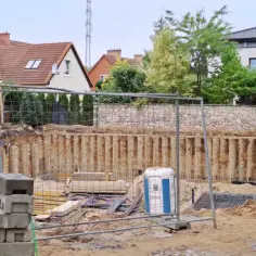 Budowa płyty fundamentowej: widok wykopu z zabezpieczeniem ścian, materiałami budowlanymi i przenośną toaletą. W tle domy i zieleń.