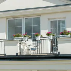 Białe okna na szarej elewacji domu, balkon z kwiatami i meblami.