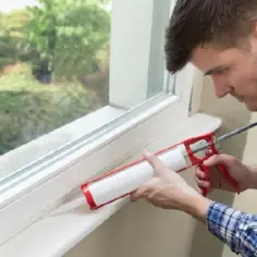 Mężczyzna uszczelnia okno plastikowe przy parapecie, używając pistoletu do silikonu.