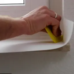 Dłoń z żółtą szpachelką przykleja białą okleinę na parapet. Poradnik: jak przykleić okleinę na parapet.