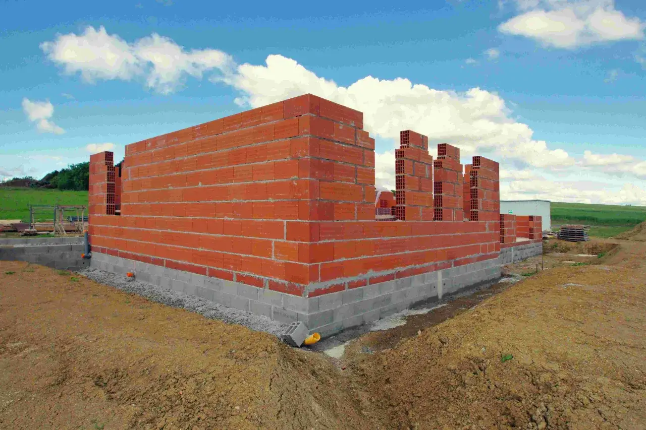 Budowa domu parterowego w toku. Wzniesione ściany z cegły na fundamencie z bloczków betonowych. W tle zielone pole i błękitne niebo z chmurami.