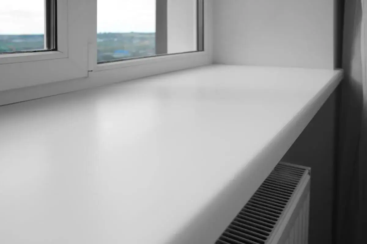 Biały, szeroki parapet okienny z tworzywa sztucznego, pod którym widać grzejnik. Czy parapet jest konieczny? Tak, dla estetyki i funkcjonalności.